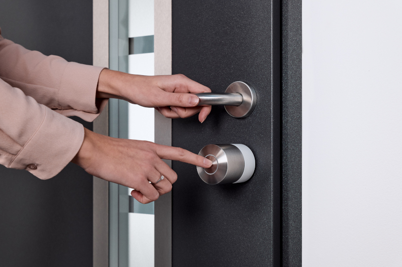 La imagen muestra una puerta moderna con un sistema de cerradura inteligente de la marca Nuki. Una persona está utilizando el dispositivo para desbloquear la puerta, lo que ilustra la funcionalidad de la domótica en el hogar.