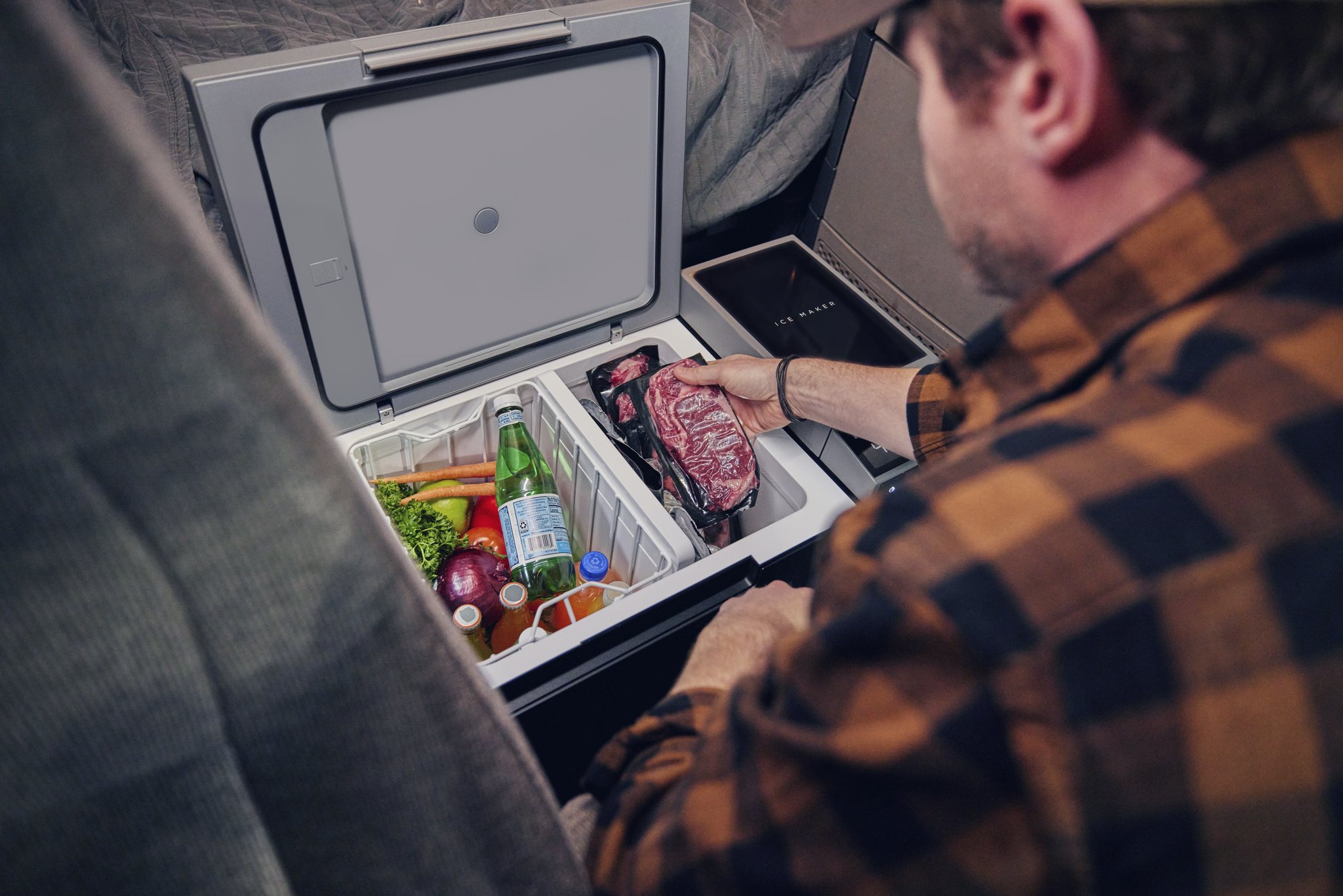 La imagen muestra una nevera portátil EcoFlow Glacier, abierta y en uso. Se puede ver a una persona colocando un paquete de carne en el compartimento, mientras que en el interior hay verduras, botellas de agua y refrescos. El ambiente es acogedor, posiblemente en un camping o una furgoneta, con colores neutros y un enfoque en la funcionalidad del producto. La nevera tiene un diseño moderno y práctico, ideal para actividades al aire libre.