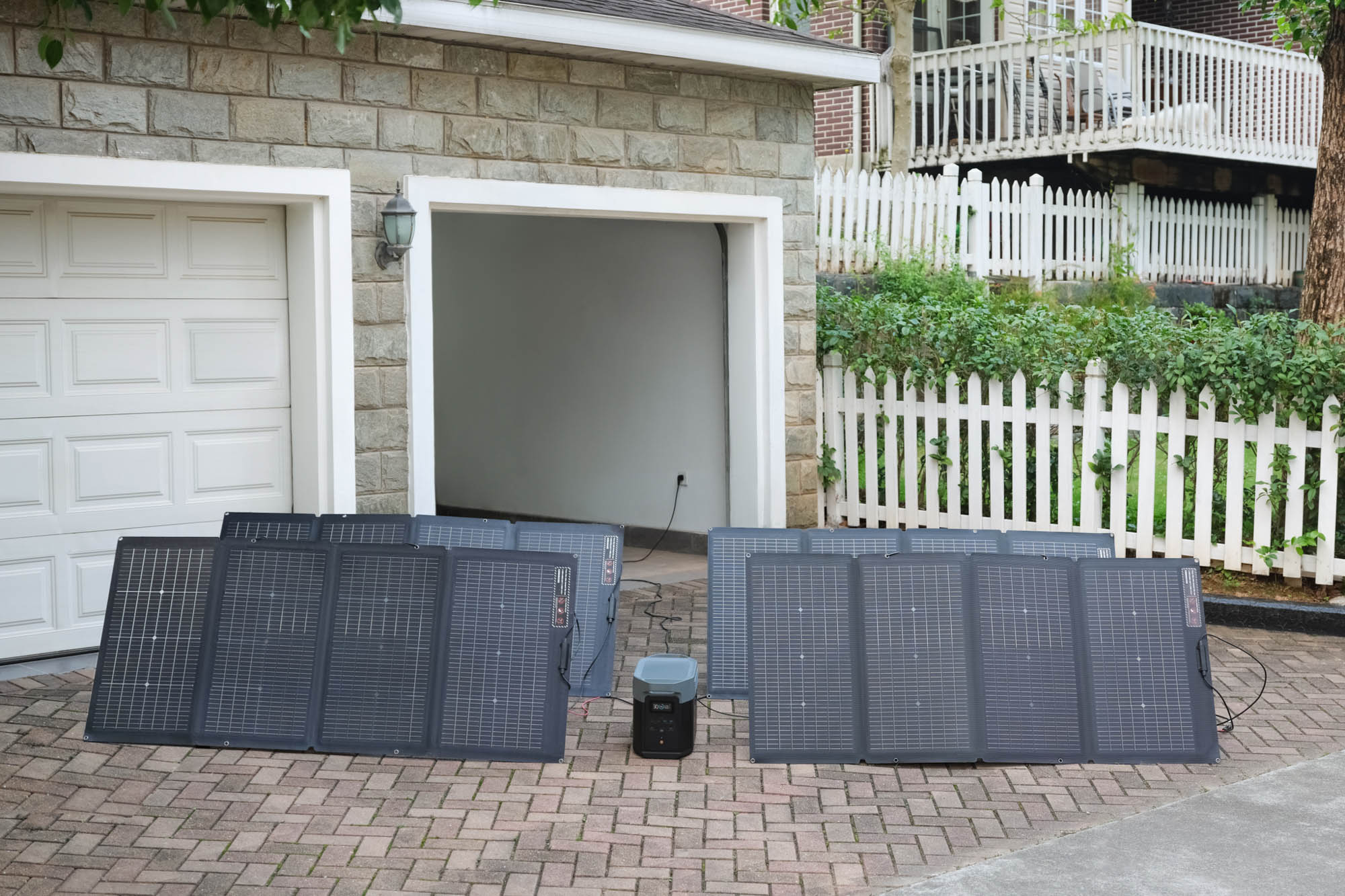 La imagen muestra un conjunto de paneles solares dispuestos en el suelo, conectados a una unidad de energía EcoFlow DELTA 2 Max. Los paneles son de color negro y están alineados en un entorno exterior, con una casa de fondo y una cerca blanca visible. La escena está iluminada por luz natural, lo que resalta los detalles de los paneles y la unidad. Este tipo de instalación es ideal para la generación de energía renovable en espacios residenciales.