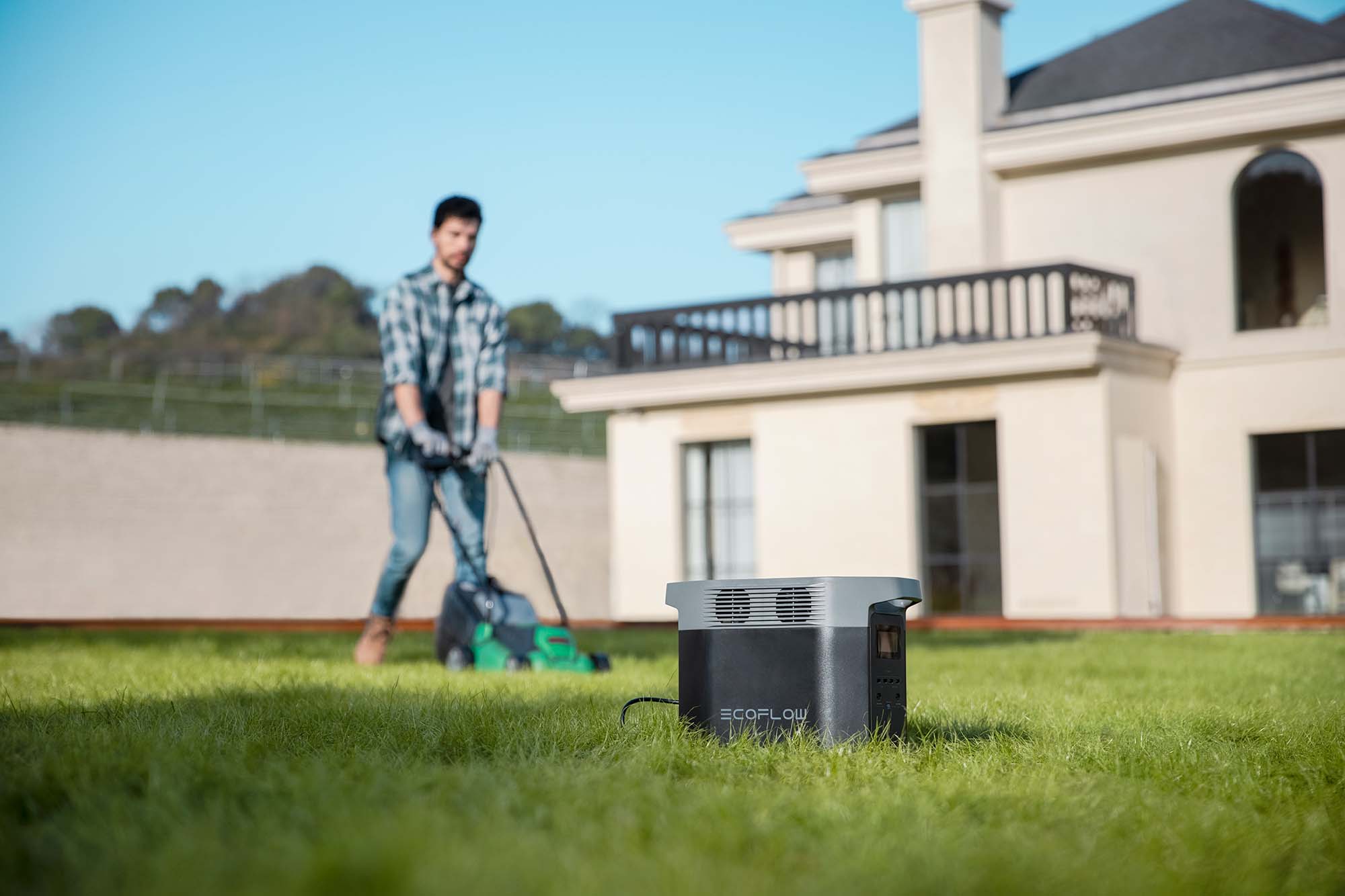 La imagen muestra un generador portátil EcoFlow DELTA 2 ubicado en un jardín bien cuidado. En el fondo, un hombre está utilizando una cortadora de césped, lo que sugiere que el generador está siendo utilizado para alimentar herramientas de jardinería. La escena es luminosa y al aire libre, con un cielo azul claro y una casa moderna al fondo. El generador tiene un diseño elegante y moderno, con colores gris y negro.