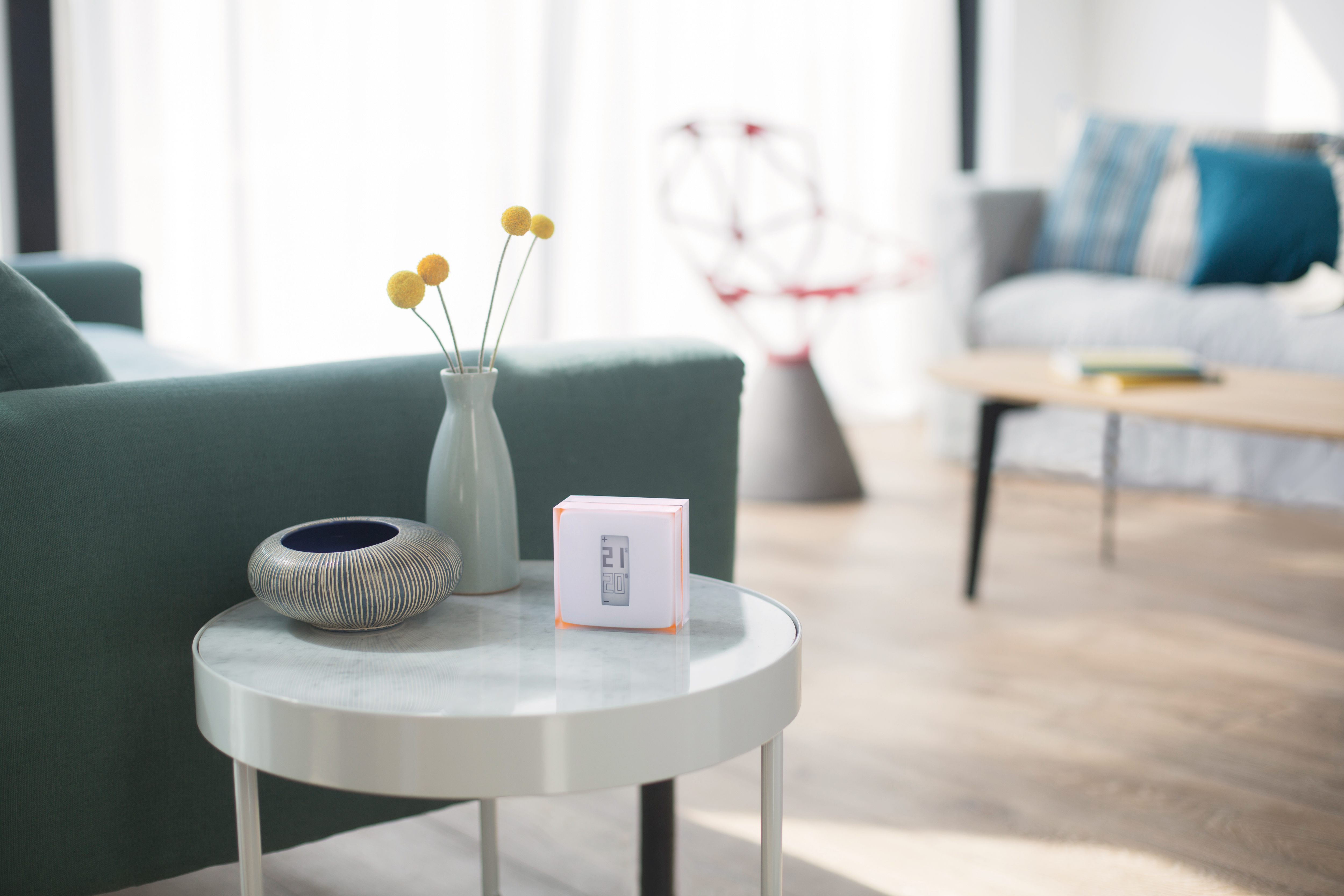 La imagen muestra un termostato de la marca Netatmo colocado sobre una mesa de centro en un ambiente moderno y acogedor. El termostato está en una caja de presentación con un diseño minimalista, destacando su forma rectangular y colores suaves. Al fondo, se puede ver un sofá verde y una decoración elegante que incluye un jarrón con flores amarillas, lo que sugiere un espacio de vida contemporáneo y bien iluminado. La composición resalta la funcionalidad del producto en un entorno doméstico.