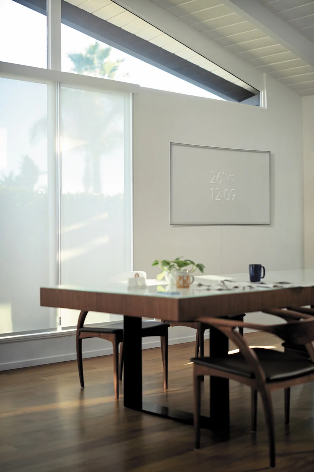 La imagen muestra un televisor Samsung QLED montado en la pared de un ambiente moderno y minimalista. El televisor presenta un diseño elegante con un marco delgado y muestra la temperatura y la hora en un formato digital. La habitación tiene una mesa de comedor de madera con sillas de diseño contemporáneo, y grandes ventanales que permiten la entrada de luz natural. Los colores predominantes son tonos claros y cálidos, creando una atmósfera acogedora.