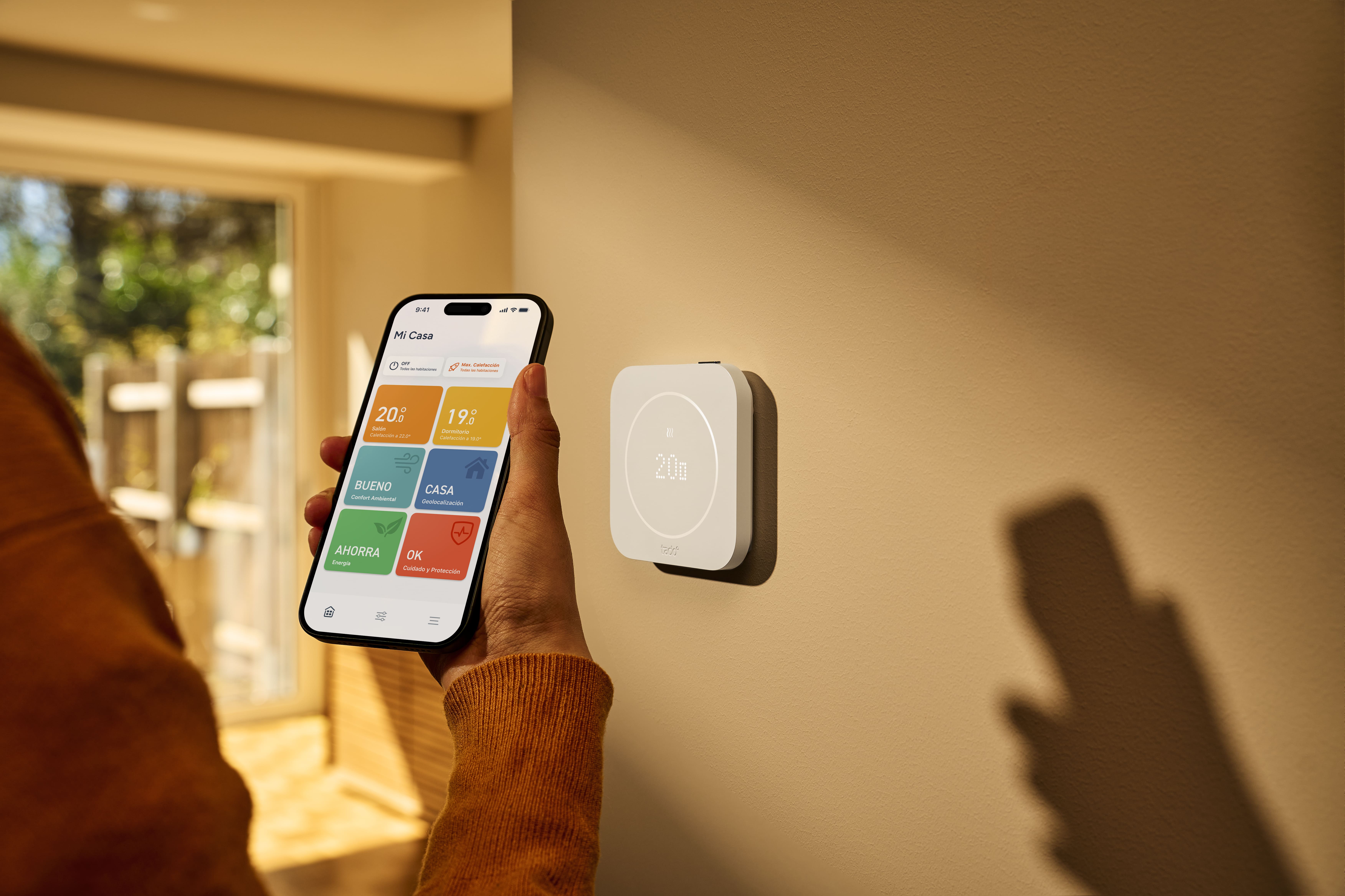 La imagen muestra un termostato inteligente montado en la pared, junto a una persona que sostiene un teléfono móvil. En la pantalla del teléfono se puede ver una aplicación que muestra información sobre el clima y el estado de la casa, indicando temperaturas y recomendaciones de ahorro energético. Este dispositivo es ideal para el control de la temperatura en el hogar de manera eficiente.